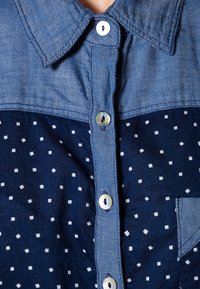 Camicia in denim con una parte superiore azzurro chiaro e una parte inferiore blu navy, caratterizzata da un motivo a quadrati bianchi; colletto classico e apertura con bottoni.