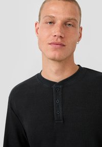 Nuori mies, jolla on lyhyet hiukset ja korvakorut, pukeutuneena mustaan urheilupaitaan, jossa on kolme nappia, katsoo suoraan eteenpäin yksinkertaista taustaa vasten.