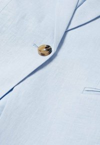 Tissu bleu clair d'un blazer avec un bouton marron marbré cousu près du revers et une petite poche visible.