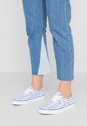 Witte canvas sneakers met meerkleurige "VANS"-tekst en hartpatronen, gecombineerd met blauwe denimspijkerbroek met een contrasterend blauw paneel en een versleten zoom.