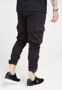 Zwarte cargojoggers met slim fit, zijzakken, geribde manchetten en textuurdetails. Gedragen met zwarte sneakers met gouden accenten.