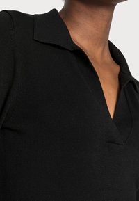 Polo shirt nero, tessuto a coste, colletto corto, scollo a V profondo, silhouette aderente, tessuto liscio e dettagli di cucitura visibili nella zona del collo.