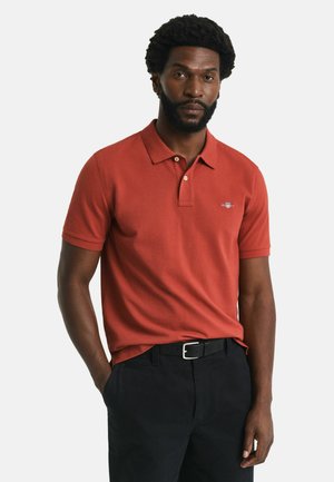 Red cotton polo shirt with a collar and three-button placket, featuring subtle branding on the chest, paired with black trousers.