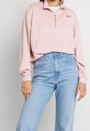 Mujer con sudadera Nike de cremallera corta de color rosa claro y vaqueros de tiro alto azul claro, posando frente a un fondo liso.