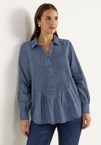 Light blue chambray shirt with a button-down collar, vertical pleats, and a flared waist. Long sleeves and textured fabric.