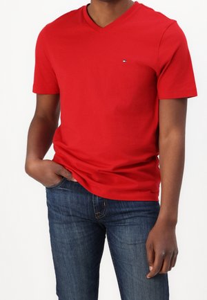 Homme portant un t-shirt rouge à manches courtes et col en V, et un jean bleu, avec la main gauche dans la poche et le bras droit détendu sur le côté.