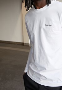 Person in einem weißen Langarmhemd von Calvin Klein und schwarzen Hosen, der drinnen steht und eine Hand in der Tasche hat.