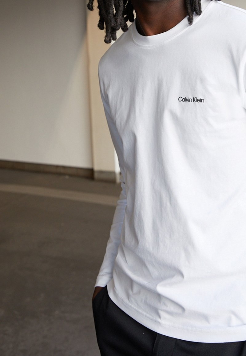 Person in einem weißen Langarmhemd von Calvin Klein und schwarzen Hosen, der drinnen steht und eine Hand in der Tasche hat.
