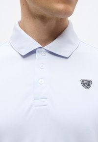 Homme portant un polo blanc à trois boutons avec un petit logo sportif noir et blanc sur le côté gauche de la poitrine.