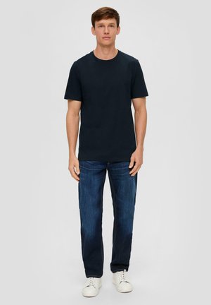 Marineblaues Baumwoll-T-Shirt mit kurzen Ärmeln und Rundhalsausschnitt, kombiniert mit blauen Jeans und weißen Sneakers, stehend vor einem neutralen Hintergrund.