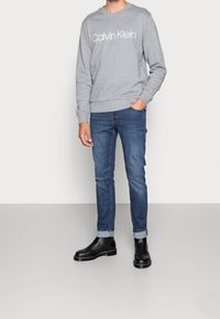 Felpa grigia con logo "Calvin Klein", jeans in denim blu e stivali Chelsea neri. Tessuto texturizzato, vestibilità slim e orli arrotolati sui jeans.
