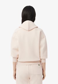 Sudadera rosa claro con una gran capucha y hombros caídos. Textura suave, puños acanalados y pequeño detalle del logo cerca del bolsillo.