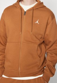 Felpa con zip marrone realizzata in tessuto morbido, con tasche frontali, cappuccio regolabile con cordino e logo Jumpman bianco sul lato sinistro del petto.