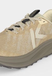 Chaussure de sport beige et beige clair avec des lacets à motifs, une tige en mesh et une semelle épaisse en caoutchouc, conçue pour un usage extérieur ou décontracté.