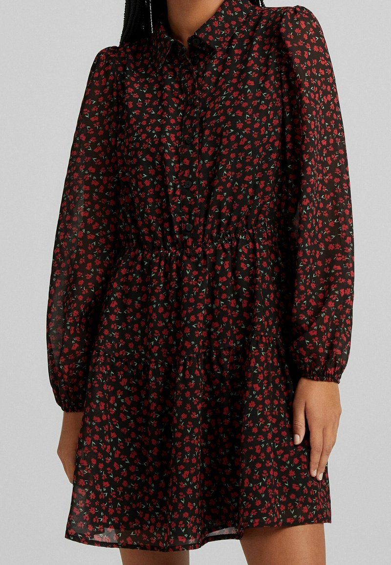 Robe florale noire avec des fleurs rouges, manches longues bouffantes, patte de boutonnage et taille élastique, avec un ourlet à couches superposées. Fabriquée en tissu léger.