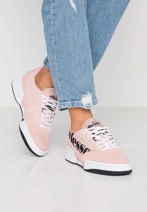 Persona que lleva vaqueros desgarrados azul claro y zapatillas rosas Ellesse con suelas blancas, de pie sobre un suelo blanco y liso.