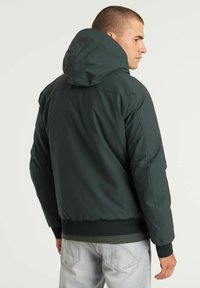 Chaqueta con capucha de color verde oscuro, con un acabado suave y mate. Presenta puños y dobladillo elásticos, así como bolsillos laterales en ángulo. Se muestra la vista posterior.