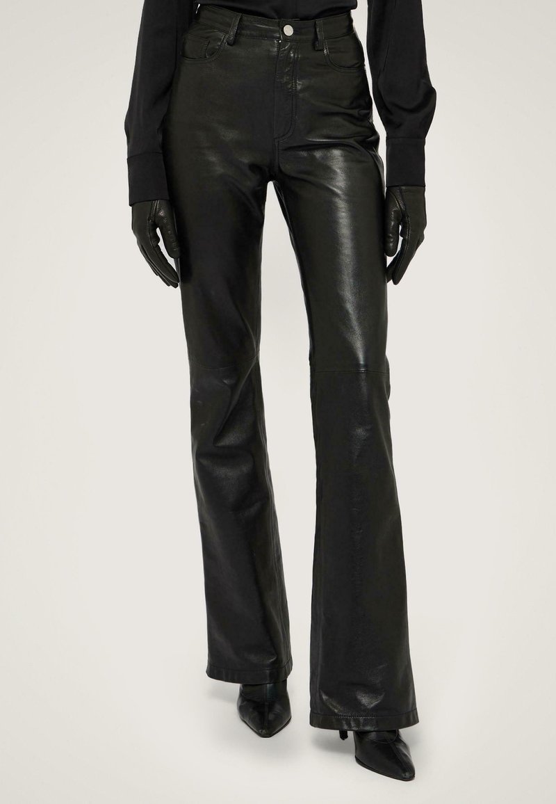 Černé kožené kalhoty s rozšířenými nohavicemi mají vysoký pas, přední kapsy a hladkou, lesklou texturu. Jsou doplněny špičatými černými podpatky.