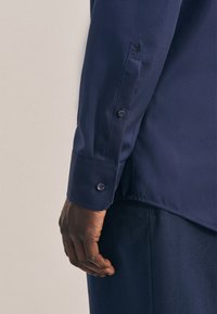 Camicia a maniche lunghe blu navy con una texture di tessuto liscia, polsini abbottonati e un delicato ricamo sul maniche. Fit sagomato visibile.