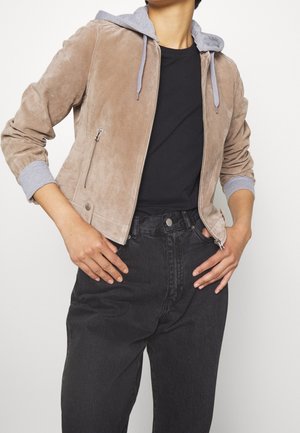 Person trägt eine beige Wildlederjacke mit grauen Kapuzenärmeln über einem schwarzen T-Shirt und hoch taillierten schwarzen Jeans, Hände auf den Hüften ruhend.