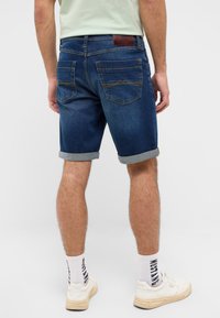 Dżinsowe szorty w ciemnym niebieskim kolorze z podwiniętym dołem, z tylnymi kieszeniami i brązową skórzaną łatą na pasie. Gładka tekstura, casualowy design.