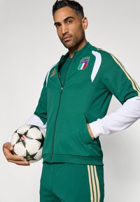 Zelený športový komplet s bielymi akcentmi a zlatými pruhmi. Obsahuje nášivku s talianskou vlajkou a zipsové vrecká, v ktorých sa nachádza futbalová lopta.