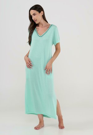 Mujer descalza con un vestido maxi suelto de color turquesa claro, de manga corta y escote en V, de pie frente a un fondo liso.