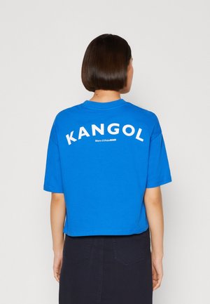 Person mit kurzen dunklen Haaren trägt ein leuchtend blaues, kurzes T-Shirt mit dem Text "KANGOL" auf dem Rücken und einen dunklen Rock.