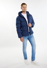 Marineblå pufferjakke med hætte, udstyret med lynlås og trykknapper, parret med lyseblå jeans og hvide sneakers.