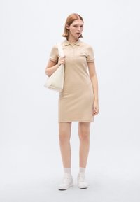 Μπεζ φόρεμα πόλο με κοντά μανίκια, γιακά και κουμπιά. Το μοντέλο κρατά μια κρεμ τσ άντα ώμου με χαλαρή μορφή. Λευκά αθλητικά παπούτσια και κάλτσες αστραγάλου ολοκληρώνουν την εμφάνιση.