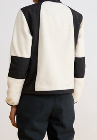 Kurtka polarowa z czarnymi i kremowymi panelami, z wysokim kołnierzem i kontrastującymi teksturami. Posiada czarne akcenty na rękawach.