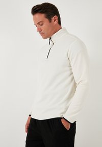 Hvid fleece-pullover med halvlukket krave, sorte detaljer på lynlåsen og ærmerne, samt en blød, tekstureret overflade.