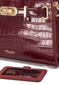 Glanzende donkerrode handtas met krokodillenpatroon en gouden hardware, Dune London-logo, en een bijpassende rechthoekige spiegel op een witte ondergrond.