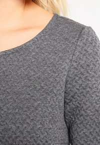 Haut gris texturé avec un motif en diamant subtil, doté d'un large col et d'un tissu lisse, offrant un ajustement confortable.