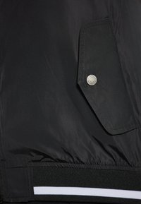 Jachetă neagră tip bomber, cu un buzunar lateral dotat cu un buton metalic, material texturat, talie elastică cu dungi albe și detalii cu nervuri.