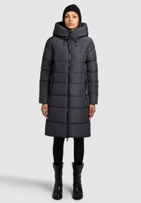 Čierny prešívaný kabát s vysokým golierom a veľkou kapucňou. Má predný zips, dve bočné vrecká a stredne dlhý strih. Hladká textúra.