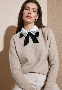 Donna con lunghi capelli castani, che indossa un maglione beige e una camicia bianca con colletto, decorata con un nastro nero e ornamenti argentati.