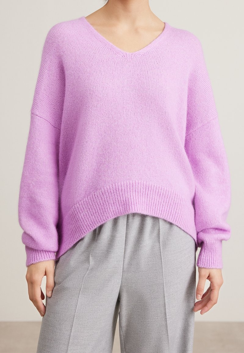 Personne portant un pull ample à col en V lavande avec des épaules tombantes et un pantalon plissé gris clair, debout les mains détendues.