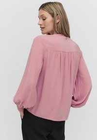 Vaaleanpunainen pitkähihaista paitaa, jossa on korkea kaulus ja puffi-hihat, takana kokoontumista ja nappikiinnitys kauluksessa.