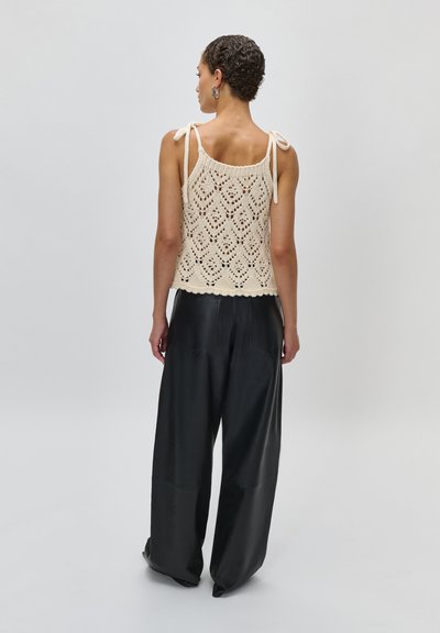 Γυναίκα με κοντά σγουρά μαλλιά φοράει μπεζ πλεκτό αμάνικο τοπ με κορδόνια στη ράχη και μαύρο παντελόνι φαρδιάς γραμμής, στέκεται μπροστά σε μια απλή φόντο.