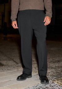 Pantaloni neri con un fit rilassato, caratterizzati da una texture liscia e pieghe sottili. Abbinati a stivali neri chunky, su una superficie in pietra.