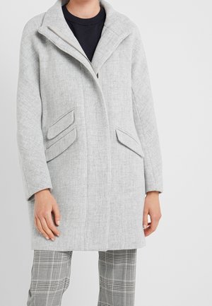 Persoon draagt een lichtgrijze jas met brede kraag, schuine klepzakken, zwarte top en grijze geruite broek tegen een effen achtergrond.