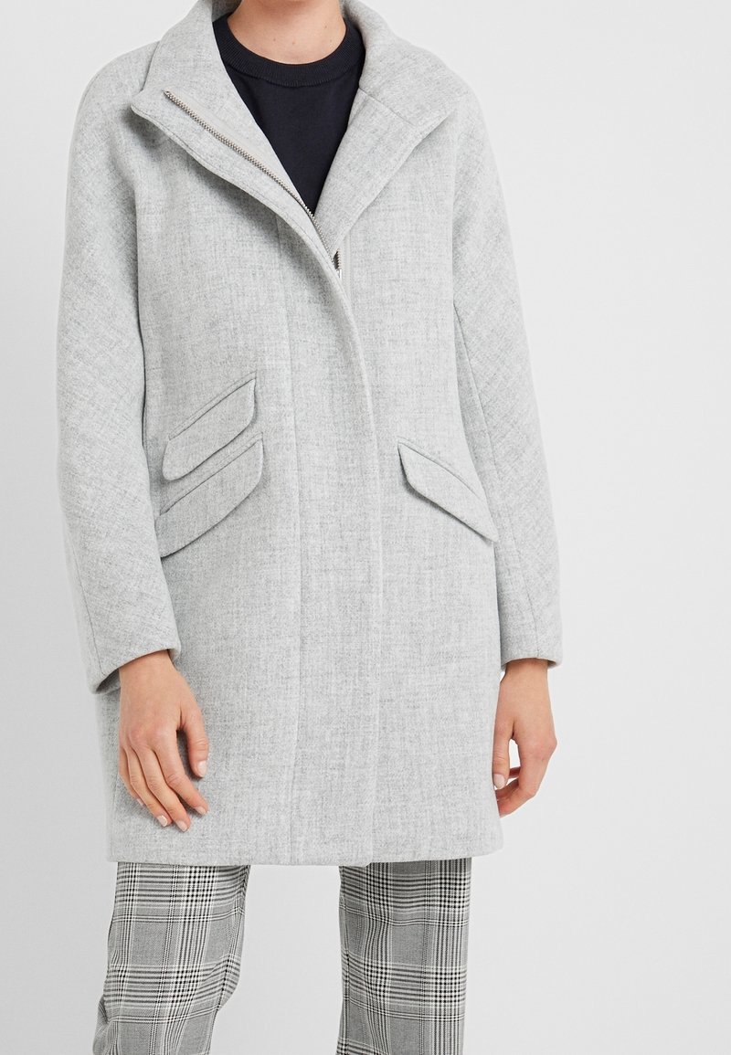 Personne portant un manteau gris clair à large col, poches à rabat inclinées, haut noir et pantalon à carreaux gris sur un fond uni.