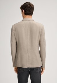 Blazer beige strutturato con silhuette sagomata, revers a punta e uno spacco singolo sul retro, abbinato a jeans scuri.