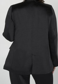 Blazer negro ajustado hecho de una tela lisa y brillante. Presenta un hombro estructurado, una abertura trasera única y un diseño minimalista sin cierres visibles.