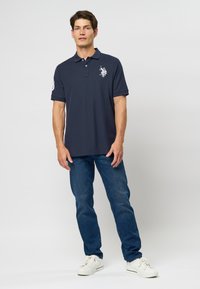 Námornícky polo tričko s golierom a krátkymi rukávmi; obsahuje biele logo na hrudi a detail s číslom na rukáve. Nosí sa s modrými džínami.