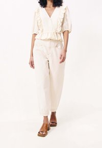 Krämfärgad blus med volanger och djup V-ringning, kombinerad med ljusbeige löst sittande byxor. Bruna sandaler med öppna tår och klack fulländar looken.