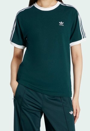 Osoba ubrana w ciemnozieloną koszulkę Adidas i pasujące spodnie z białymi paskami i logo, stojąca z jedną ręką w kieszeni.