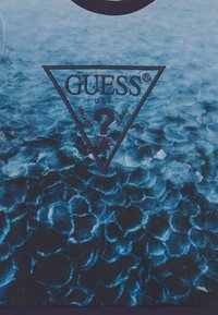 Felpa con motivo a gradiente blu, caratterizzata da un logo triangolare "GUESS" in blu scuro, con il testo "WASHED JEANS" e un design con punto interrogativo.