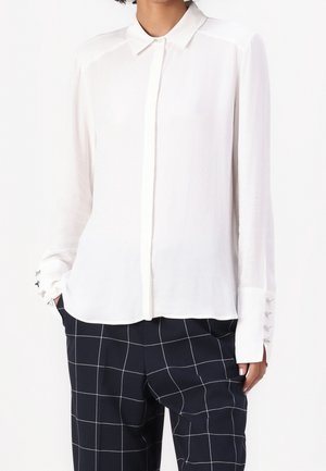 Kobieta ubrana w białą bluzkę z długim rękawem z zapinanymi mankietami i czarne spodnie w kratę, dłonie w kieszeniach, na jednolitym tle.